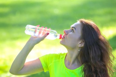 Uzun saçlı bir kadın güneşli bir parkta spor yaparken şişeden su içer. Parlak yeşil çimen, spor programının hareketli atmosferini arttırır..