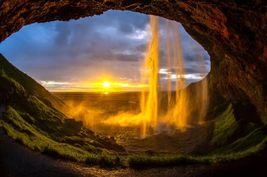 Altın güneş ışığı mağaradan çağlayan şelaleyi aydınlatır nefes kesen bir günbatımı gibi gökyüzünü renklendirir. Bereketli yeşil çevre bu İzlanda manzarasının doğal güzelliğini artırır..