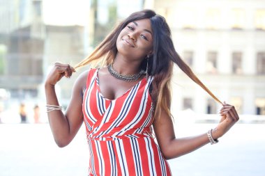 Kendine güvenen Afrikalı kadın dışarıda duruyor, gülümsüyor ve uzun saçlarını şekillendirirken anın tadını çıkarıyor. Parlak elbisesi ve neşeli tavırları güneşli bir günün özünü yansıtıyor..