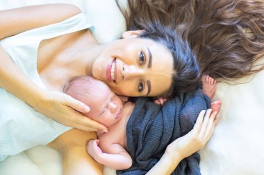 A mother is gently cradling her newborn baby while lying on soft bedding. Sunlight softly illuminates the cozy indoor space, enhancing the tender moment shared between them.