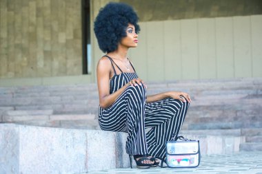 Seated on a stone step, a woman with an afro hairstyle showcases her fashion sense in a striped jumpsuit. The outdoor setting highlights her confident demeanor and modern style.