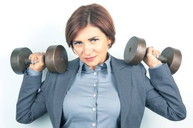 A confident woman in a tailored suit lifts dumbbells overhead, showcasing strength and determination. Set against a simple background, the scene reflects a blend of professionalism and fitness.