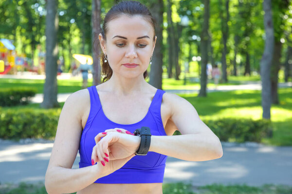 Женщина в ярко-синем спортивном топе следит за своей статистикой тренировок в фитнес-часах в пышном парке. Окруженная зелеными деревьями и пешеходными дорожками, она кажется сосредоточенной и решительной. Солнце светит, создавая яркую атмосферу, когда она участвует в