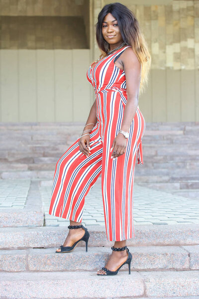 An African woman stands gracefully on steps, showcasing a stylish striped jumpsuit. Her poised demeanor highlights her confidence while she enjoys a sunny day in an urban environment.