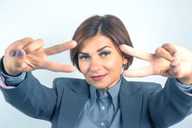 A woman with short brown hair and blue eyes smiles cheerfully while making peace signs with her fingers.