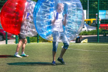 Two players participate in bubble soccer, wearing inflatable spheres while enjoying a vibrant day on an artificial turf field. The atmosphere is lively with bright colors and energetic movement.