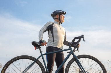 Bisiklet kıyafetli bir adam bisikletiyle duruyor, parlak mavi bir gökyüzünün altında ufka bakıyor. Sahne, doğada bisikletle ilişkili macera ve özgürlük duygusunu yakalar..