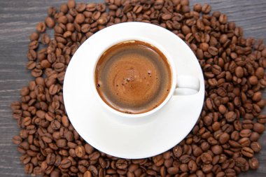 Yukardan bakıldığında kremalı köpüklü küçük beyaz bir fincan espresso sergileniyor. Bir fincan çay tabağının üzerinde ve koyu kavrulmuş kahve çekirdekleri ile çevrelenmiş..