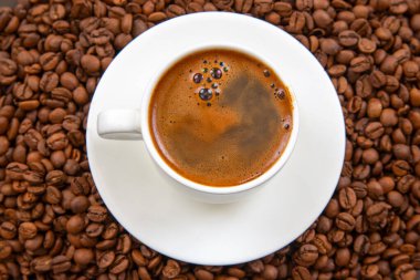 Koyu kahve ve hafif kremayla dolu beyaz bir espresso fincanı, tepeden tırnağa kapalı bir perspektiften yakalanmış, kavrulmuş kahve çekirdekleri ile çevrili, uyumlu bir fincan üzerinde oturur.