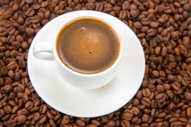 Beyaz seramik bir fincanda köpüklü krema ile zengin, koyu espresso. Dağılmış kahve çekirdekleri arasında zarif bir şekilde bir fincan üzerine yerleştirilip sıcak, davetkar bir kahve sahnesi yaratıyor.