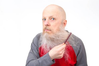 Uzun beyaz sakallı kel yaşlı bir adam kırmızı bir tarak ile sakalını tarıyor, kırmızı ve gri atletik bir gömlek giyiyor, düz beyaz bir arka plan üzerine kurulmuş, mizahi bir tımar anı sergiliyor.