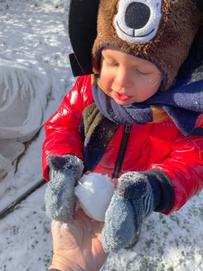 Kırmızı ceketli, eldivenli ve kalın şapkalı küçük çocuk kartopu alır. Kışın güneşli bir gün. Kar ve mevsimler. - Evet. Yüksek kalite fotoğraf