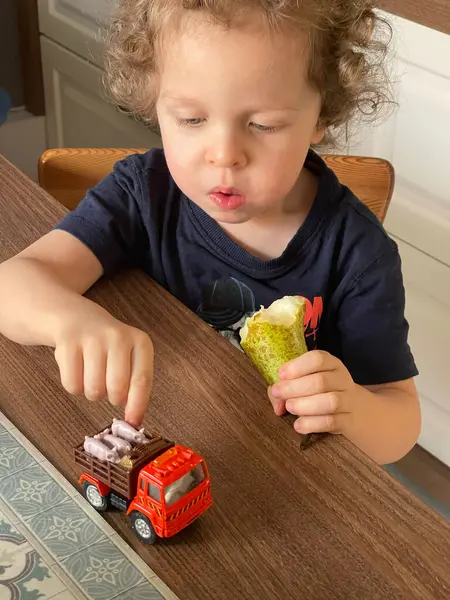 Küçük kıvırcık saçlı sarışın çocuk armut yiyor ve biri de tahta masada arabasıyla oynuyor. Çocuklar için geliştirme oyunları. - Evet. Yüksek kalite fotoğraf