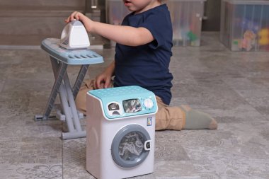 Küçük çocuk evde yerde oyuncak ütü ve çamaşır makinesiyle oynuyor. Temizlik, ev işleri, çocuklar ve eğitim oyunları. - Evet. Yüksek kalite fotoğraf