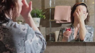 50 'li yaşlarda banyoda bornoz ve havlu giyen yaşlı bir kadın aynaya bakarken yaşlanmama önleyici krem sürüyor. Yüz bakımı. Günlük güzellik tedavileri. Yüksek kalite 4k görüntü