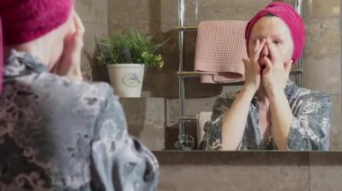 50 yaşında banyoda bornozlu yaşlı bir kadın yüz masajı yapıyor, yaşlanmayı önleyici kremler sürüyor, aynaya bakıyor. Yüz bakımı. Günlük güzellik tedavileri. Yüksek kalite 4k görüntü