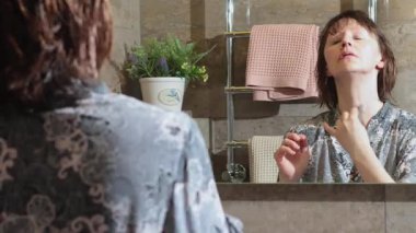 50 yaşında, banyoda bornozlu yaşlı bir kadın, aynaya bakarak boynuna agate kazıyıcı ile guasha masajı yapıyor. Yüz bakımı. Günlük güzellik spa tedavileri. Yüksek kalite 4k görüntü