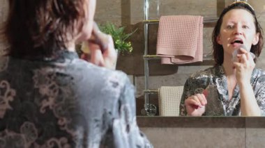 50 yaşında, banyoda bornozlu yaşlı bir kadın aynaya bakarak yüz masajı yapıyor. Yüz bakımı. Günlük güzellik spa tedavileri. Yüksek kalite 4k görüntü
