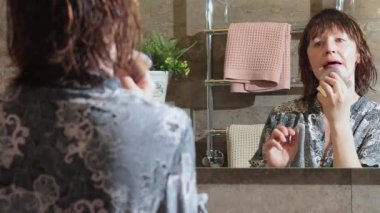50 yaşında, banyoda bornozlu yaşlı bir kadın aynaya bakarak yüz masajı yapıyor. Yüz bakımı. Günlük güzellik spa tedavileri. Yüksek kalite 4k görüntü