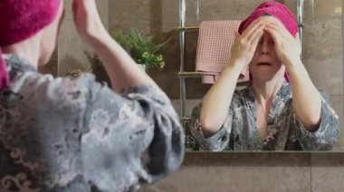 50 yaşında banyoda bornozlu yaşlı bir kadın yüz masajı yapıyor, yaşlanmayı önleyici kremler sürüyor, aynaya bakıyor. Yüz bakımı. Günlük güzellik tedavileri. Yüksek kalite 4k görüntü