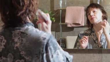 50 yaşında, banyoda bornozlu yaşlı bir kadın, aynaya bakarak boynuna agate kazıyıcı ile guasha masajı yapıyor. Yüz bakımı. Günlük güzellik spa tedavileri. Yüksek kalite 4k görüntü
