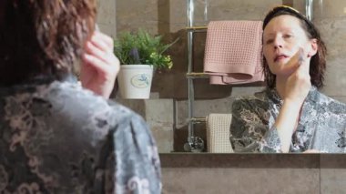 50 yaşında, banyoda bornozlu yaşlı bir kadın aynaya bakarak yüz masajı yapıyor. Yüz bakımı. Günlük güzellik spa tedavileri. Yüksek kalite 4k görüntü