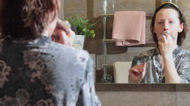 50 yaşında, banyoda bornozlu yaşlı bir kadın aynaya bakarak yüz masajı yapıyor. Yüz bakımı. Günlük güzellik spa tedavileri. Yüksek kalite 4k görüntü