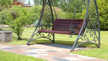 Parkın yanındaki kiremit yola paraşütle demir salıncak döşemiş. Yazın güneşli bir günde yeşil çimenler, çalılar ve ağaçlar. Tasarım için arka plan. Ekoloji ve doğa. Peyzaj tasarımı. Yüksek kalite fotoğraf