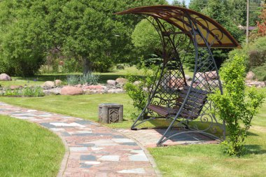 Parkın yanındaki kiremit yola paraşütle demir salıncak döşemiş. Yazın güneşli bir günde yeşil çimenler, çalılar ve ağaçlar. Tasarım için arka plan. Ekoloji ve doğa. Peyzaj tasarımı. Yüksek kalite fotoğraf