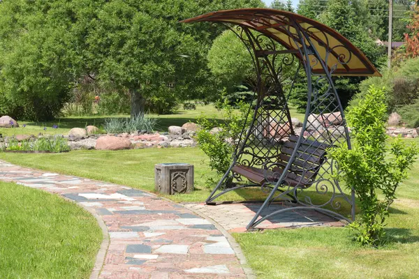 Parkın yanındaki kiremit yola paraşütle demir salıncak döşemiş. Yazın güneşli bir günde yeşil çimenler, çalılar ve ağaçlar. Tasarım için arka plan. Ekoloji ve doğa. Peyzaj tasarımı. Yüksek kalite fotoğraf