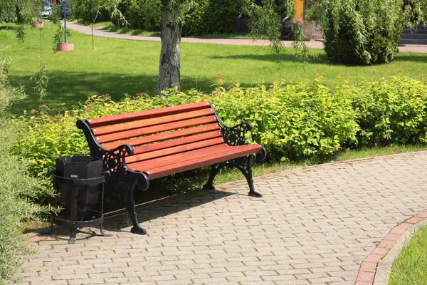 Tahta bank, parktaki fayans yolunun yanında işlenmiş demir çöp kutusu. Yazın güneşli bir günde yeşil çimenler, çalılar ve ağaçlar. Tasarım için arka plan. Ekoloji ve doğa. Peyzaj tasarımı. - Evet. Yüksek kalite fotoğraf