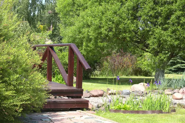 Kuru nehrin üzerindeki ahşap köprü ve parkta taşlar. Yazın güneşli bir günde yeşil çimenler, çalılar ve ağaçlar. Tasarım için arka plan. Ekoloji ve doğa. Peyzaj tasarımı. Yüksek kalite fotoğraf