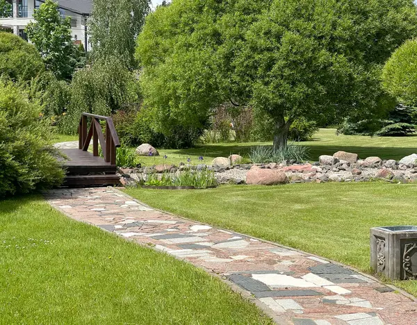 Nehrin üzerinde tahta köprü ve parkta taşlar. Yazın güneşli bir günde yeşil çimenler, çalılar ve ağaçlar. Tasarım için arka plan. Ekoloji ve doğa. Peyzaj tasarımı. Yüksek kalite fotoğraf