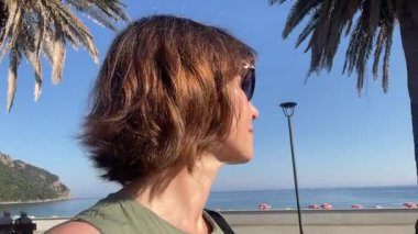 Yazın deniz kıyısında yürüyen güneş gözlüklü bir kadın. Seyahat, modern yaşam tarzı. Doğa ve mavi gökyüzü, dinlenme ve rahatlama. Yüksek kalite 4k görüntü