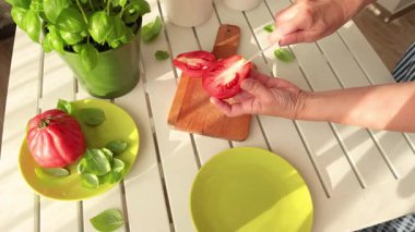 Kadın domatesi seramik bıçakla kesti. Evde sebzeler ve bitkiler var. Çömlekte fesleğen. Sağlıklı beslenme. Çevre dostu ürünler. Akdeniz diyeti. Yüksek kalite 4k görüntü