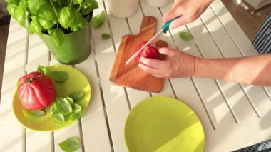 Kadın seramik bıçakla pembe domatesi ikiye böldü. Evde sebzeler ve bitkiler var. Çömlekte fesleğen. Sağlıklı beslenme. Çevre dostu ürünler. Akdeniz diyeti. Yüksek kalite 4k görüntü