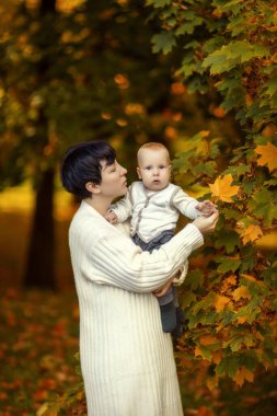 Mom in a light sweater with a small child in her arms against the background of golden autumn