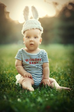 Gün batımında yeşil çimenlerin üzerinde oturan tavşan kulaklı tatlı erkek bebek.