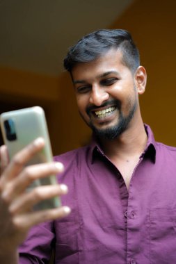  Yüzünde tebessüm olan yakışıklı bir adam elinde telefonuyla dikiliyor ve selfie çekiyor.