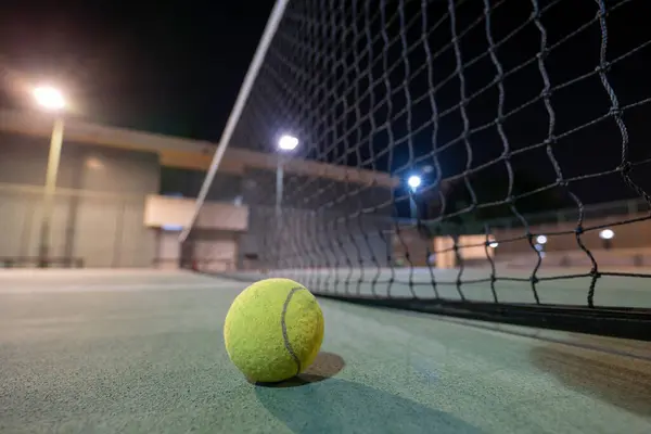 Tennis Ball Under the Lights. High quality photo tennis ball sitting on the center of a tennis court at night. The court is illuminated by bright floodlights, casting long shadows across the green