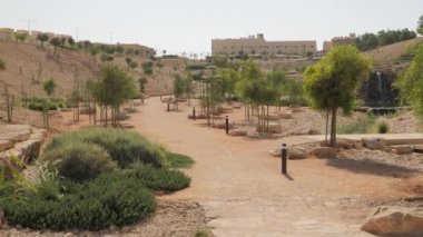 Güneşli bir günde, bir park yemyeşil yapraklarla süslenmiş uzun ağaçlarla doludur. Çakıl taşlarından yapılmış ve ağaçlar ve çalılarla çevrili dolambaçlı bir patika parkın içinden geçiyor. Bu sakin sahne