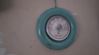 Görüntü bir duvar termometresinin yakın çekimi. Yuvarlak ve metalden yapılmış. Termometrenin şu anki sıcaklığı gösteren kırmızı bir iğnesi var. Sıcaklık Celsius dereceleriyle ölçülür.