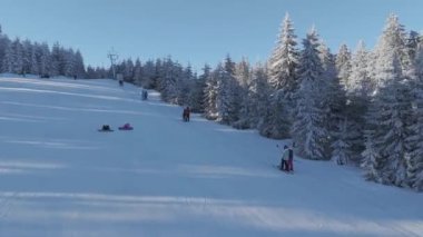 Kuş bakışı bakış açısından kayakçılar kayak asansörüne hevesle biniyorlar, rengarenk giysileri aşağıdaki karlı manzarayla çelişiyor. Asansör nazikçe yükselir, onları kuleye doğru taşır.