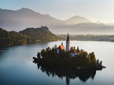 İHA uçuşu. Slovenya. Bled Gölü. Gün doğumu. Gökyüzünün parlak mavisini yansıtan berrak bir göl. Yeşiller yemyeşil dağlar turkuaz suyun etrafını sarmış ve yamaçlarında parlak noktalar var.