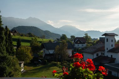 İHA uçuşu. Slovenya. Köy yemyeşil çayırlar ve yuvarlanan tepelerle çevrili bir vadiye kurulmuştur. Arka planda zirveler görkemli bir şekilde yükselir, tepeleri açık mavi gökyüzünü delip geçer.. 