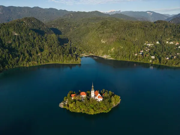 İHA uçuşu. Slovenya. Bled Gölü. Gökyüzünün parlak mavisini yansıtan berrak bir göl. Yeşiller yemyeşil dağlar turkuaz suyun etrafını sarmış ve yamaçlarında parlak yeşil noktalar var.