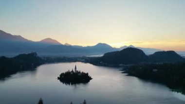 İHA uçuşu. Slovenya. Bled Gölü. Gün doğumu. Gökyüzünün parlak mavisini yansıtan berrak bir göl. Yeşiller yemyeşil dağlar turkuaz suyun etrafını sarmış ve yamaçlarında parlak noktalar var.