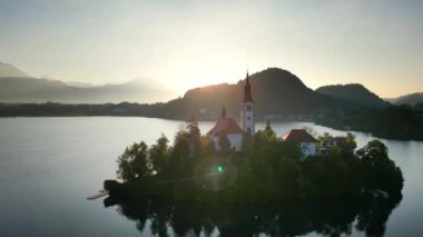 İHA uçuşu. Slovenya. Bled Gölü. Gün doğumu. Gökyüzünün parlak mavisini yansıtan berrak bir göl. Yeşiller yemyeşil dağlar turkuaz suyun etrafını sarmış ve yamaçlarında parlak noktalar var.