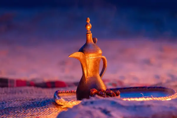 Günbatımında tarihleri olan Arap Dallah Kahve Sahası. Geleneksel bir pirinç dallah kahve demliği, bir tepsi hurma eşliğinde çöl günbatımının altın parıltısında zarif bir şekilde oturur. Bu zamansız sembol