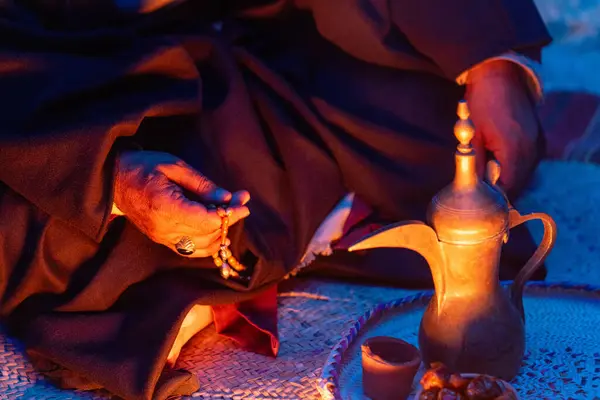 Suudi Arabistan 'da geleneksel Bedevi Konukseverliği. Bedevi bir adamın çölde örülmüş bir halıya oturmuş elinde dua boncukları ve yanında geleneksel bir Arap kahve cezvesi olan Dallah ve